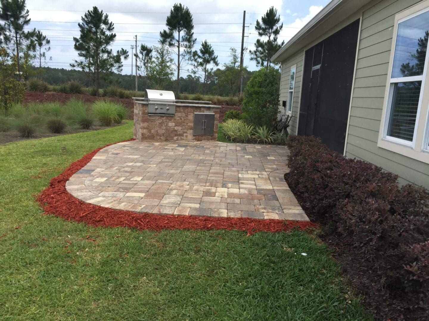 Patio area in North East Florida with brick paving and an outdoor grill, bordered by red mulch and shrubbery next to a building.