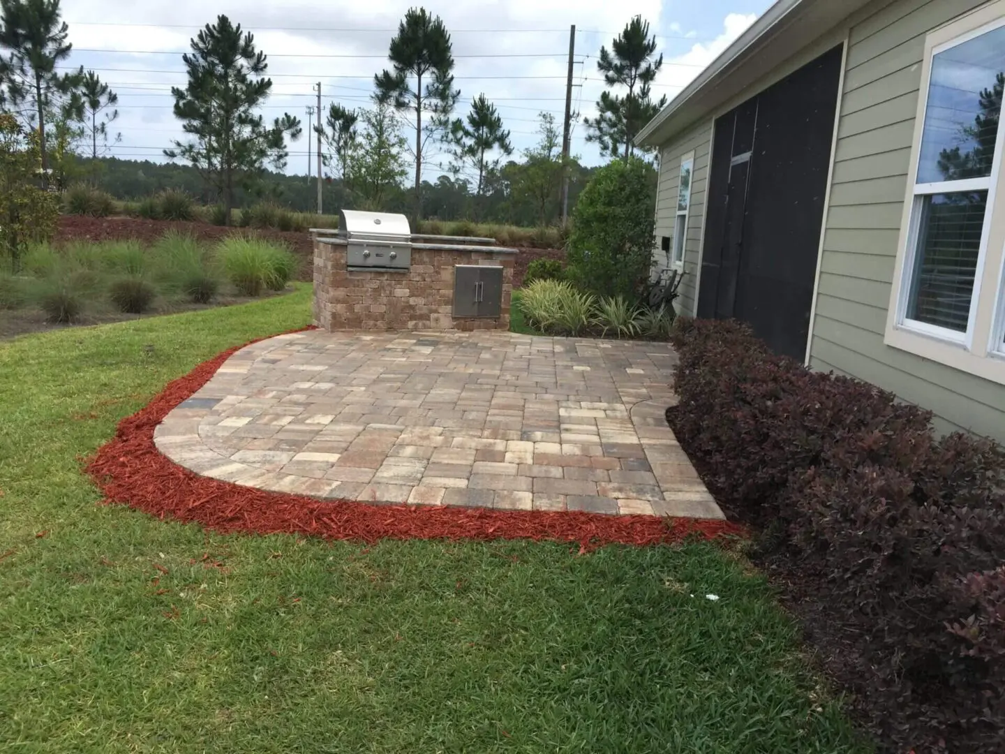 Paved patio area with an integrated grill beside a house with fresh red mulch and shrubbery.