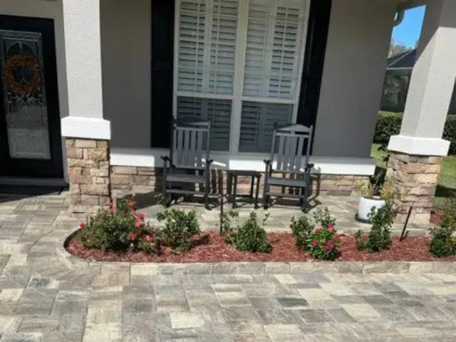Two chairs and a small table on a paved patio area with surrounding flowerbeds in front of a house.